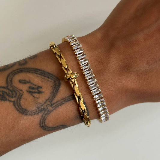 Bracelet Kezia doré serti de zircons rectangulaires porté au poignet avec un autre bracelet doré.