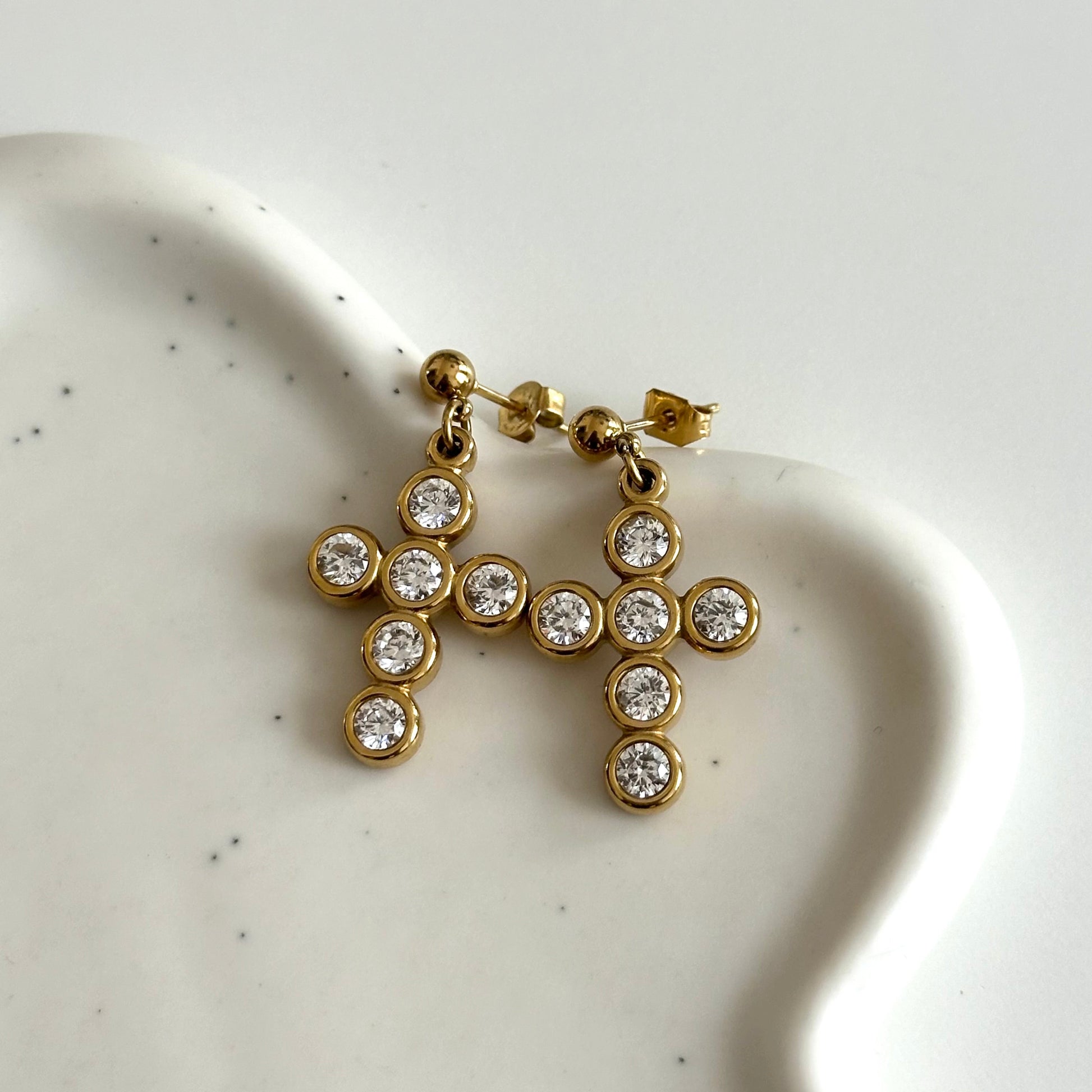Boucles d’oreilles Livia dorées en acier inoxydable PVD or 18K avec croix serties de zircons, posées sur support blanc.
