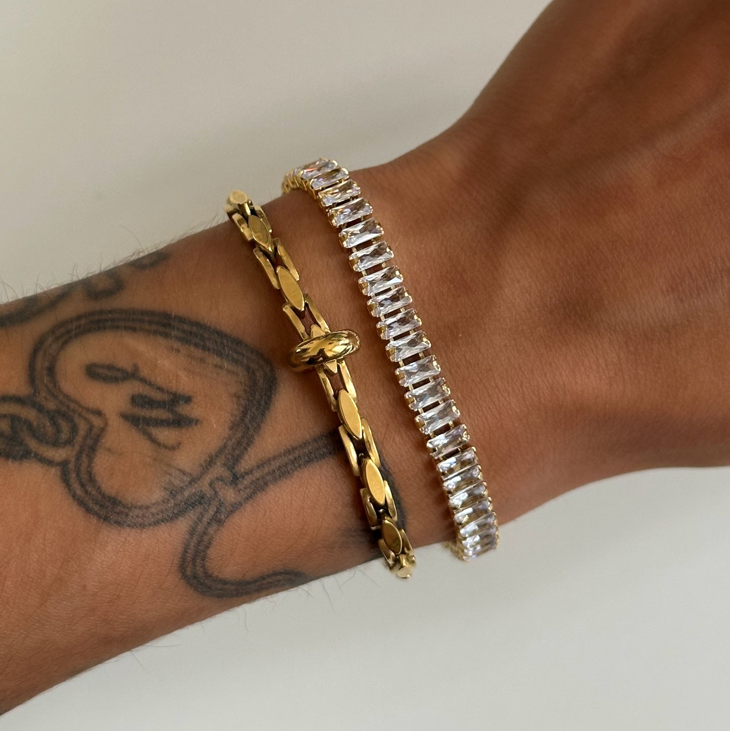 Bracelet Kezia doré serti de zircons rectangulaires porté au poignet avec un autre bracelet doré.