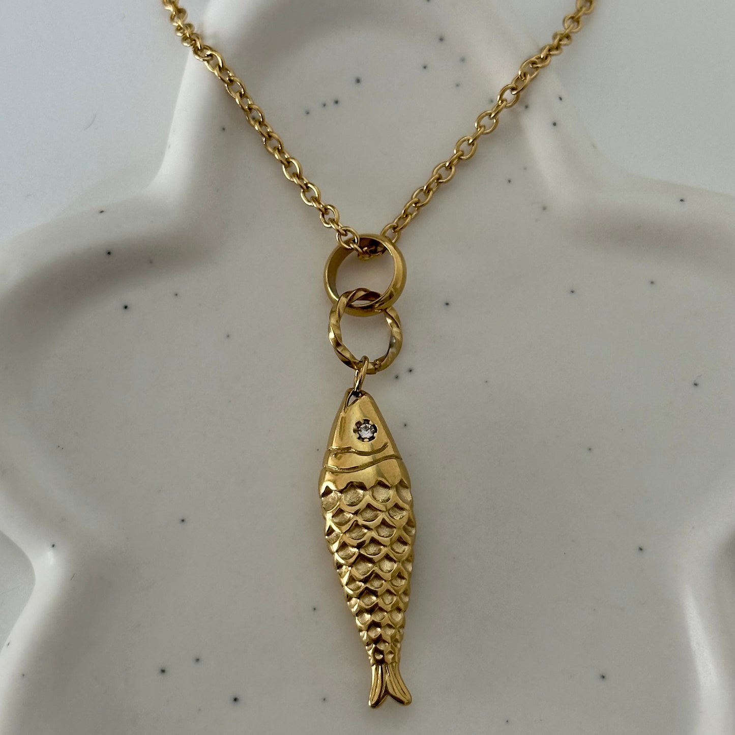Collier Ariel en acier inoxydable doré avec pendentif poisson texturé présenté sur support.