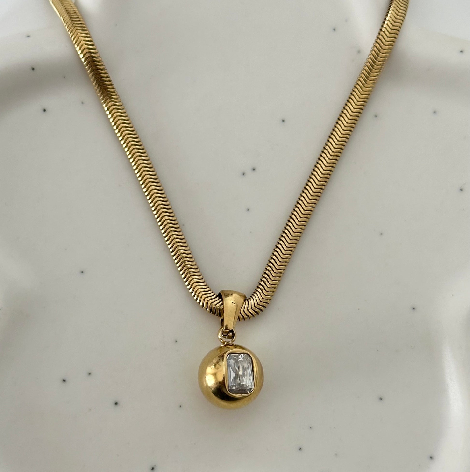 Collier Cielle en acier inoxydable doré avec pendentif boule et zircon rectangulaire présenté sur support.